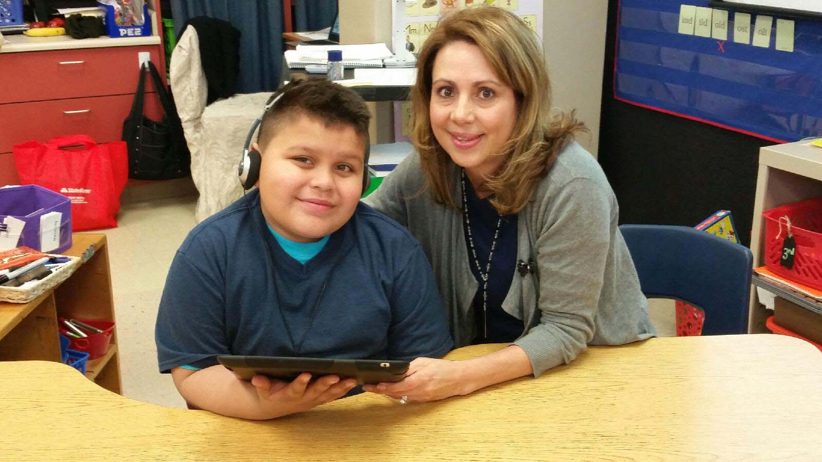Mrs. Reyes sitting with a student in class. He has headphones on and is reading a tablet.